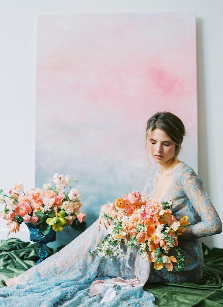 At Floral Design Studio, a woman in a blue lace gown sits beside beautiful floral arrangements, with a large pastel abstract painting in the background.