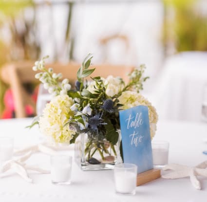 Floral Design Studio's touch shines in the event's table setting, adorned with a centerpiece, blue 'table one' card, votive candles, and white linens.