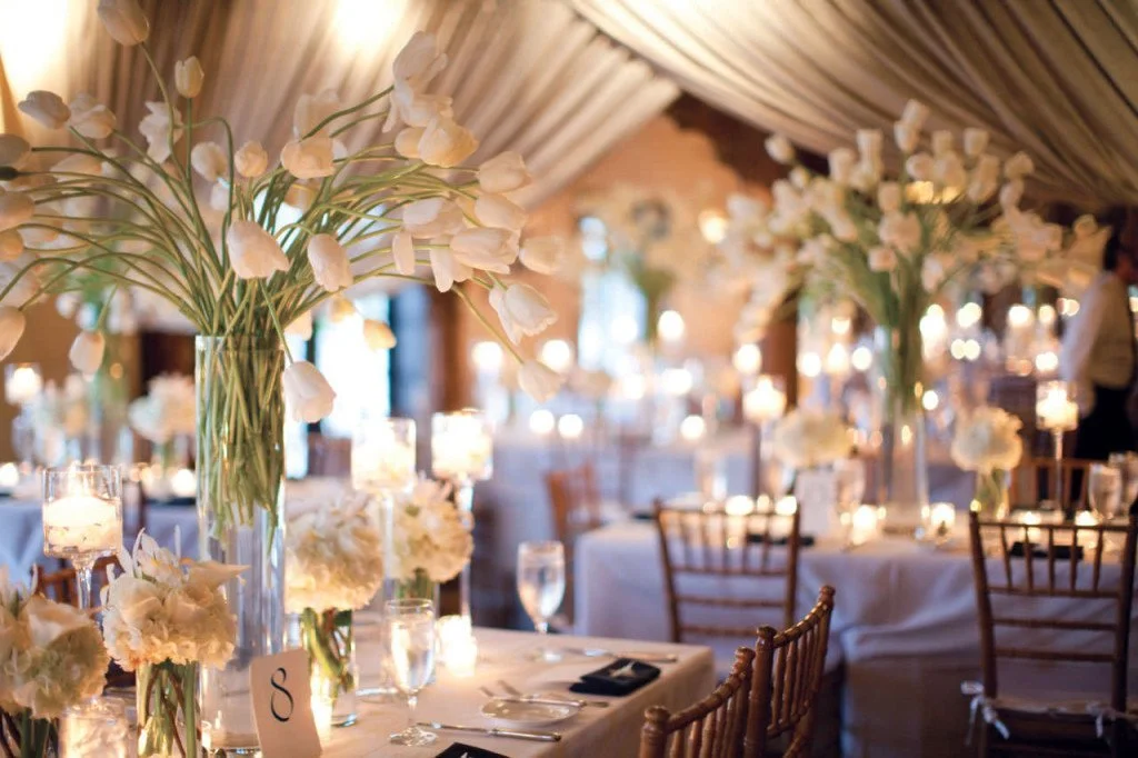 Experience the enchantment of an elegant wedding reception, artfully decorated with pristine white tulips and flickering candles, meticulously crafted by Blooming Events Florist.
