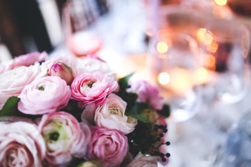 A close-up view of a floral design studio with pink and white ranunculus flowers, surrounded by soft lighting and blurry table setting elements in the background.